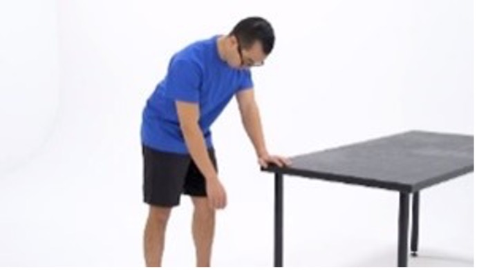 A man in a blue shirt uses the corner of a black table for support. The man is holding his arm loose at the shoulder as he begins an arm circle exercise to improve his mobility.