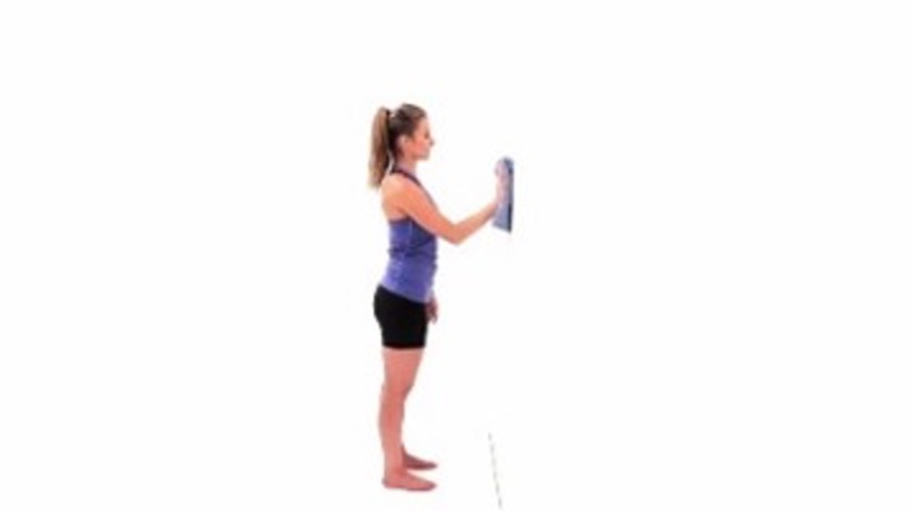 A woman in a blue tank top and black bike shorts stands with her feet shoulder-width apart, facing a white wall. Her right arm is extended against the wall, above eye level. She is holding a grey towel that she slid up the wall.