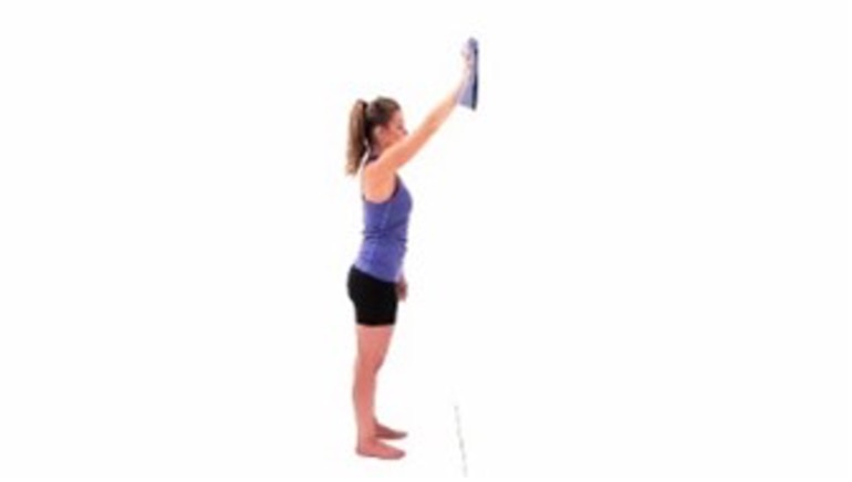 A woman in a blue tank top and black bike shorts stands with her feet shoulder-width apart, facing a white wall. Her right arm is extended against the wall, above eye level. She is holding a grey towel that she slid up the wall.