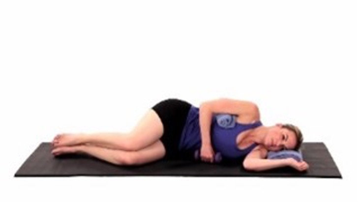 A person is shown lying on her side on a black yoga mat, wearing black shorts and a blue tank top. Her knees are bent, and her arm is resting across her body.