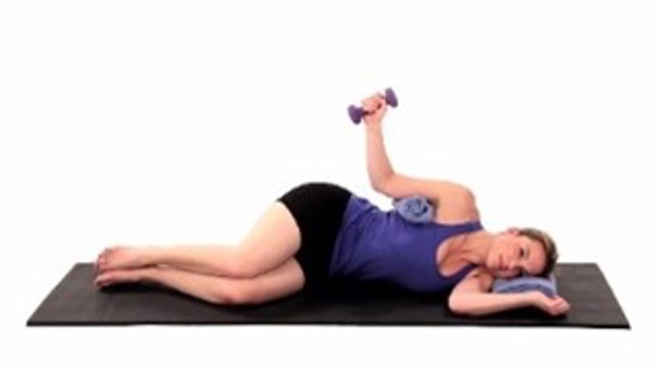 A person in a blue tank top and black shorts is on her side on a black yoga mat with a blue cushion under her head; her knees are bent. She is extending a small dumbbell up and out with her bent arm.