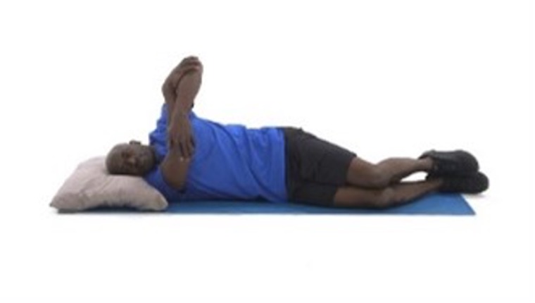 A man is wearing a blue shirt and black shorts with sneakers. He’s on his side, lying on a yoga mat with a grey pillow, as he stretches a bent arm to exercise his shoulder.