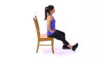 An exercise illustration depicts a brunette woman with a ponytail, wearing a bright blue tank top, black capri-length leggings, and black sneakers. The woman sits on the edge of a brown wooden chair with her back straight, hands on her hips, and one leg bent. Her other leg is extended forward with the heel resting on the floor. 